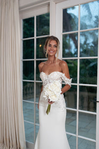 Bride Holding a White Rose Bouquet wearing Ivory White Off The Shoulder Wedding Gown with Semi Sheer Corset Style Bodice, Sweetheart Off Shoulder Neckline, Lace Appliques, Fit and Flare Silhouette and Moderate Train