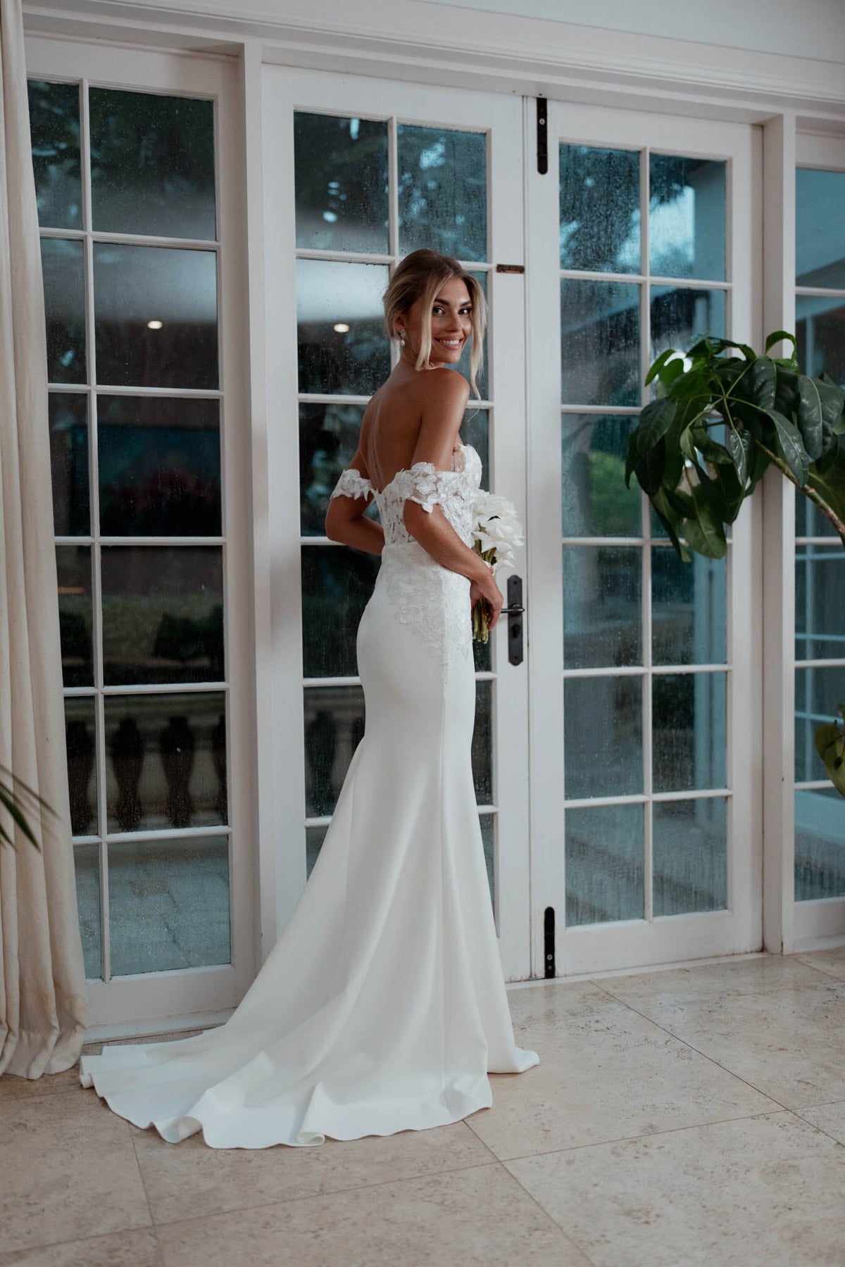 Bride Holding a White Rose Bouquet wearing Ivory White Off The Shoulder Wedding Gown with Semi Sheer Corset Style Bodice, Sweetheart Off Shoulder Neckline, Lace Appliques, Fit and Flare Silhouette and Moderate Train