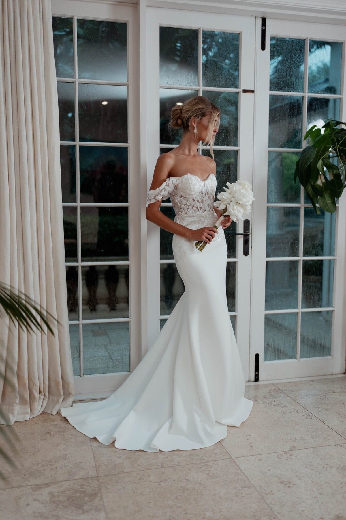 Bride Holding a White Rose Bouquet wearing Ivory White Off The Shoulder Wedding Gown with Semi Sheer Corset Style Bodice, Sweetheart Off Shoulder Neckline, Lace Appliques, Fit and Flare Silhouette and Moderate Train